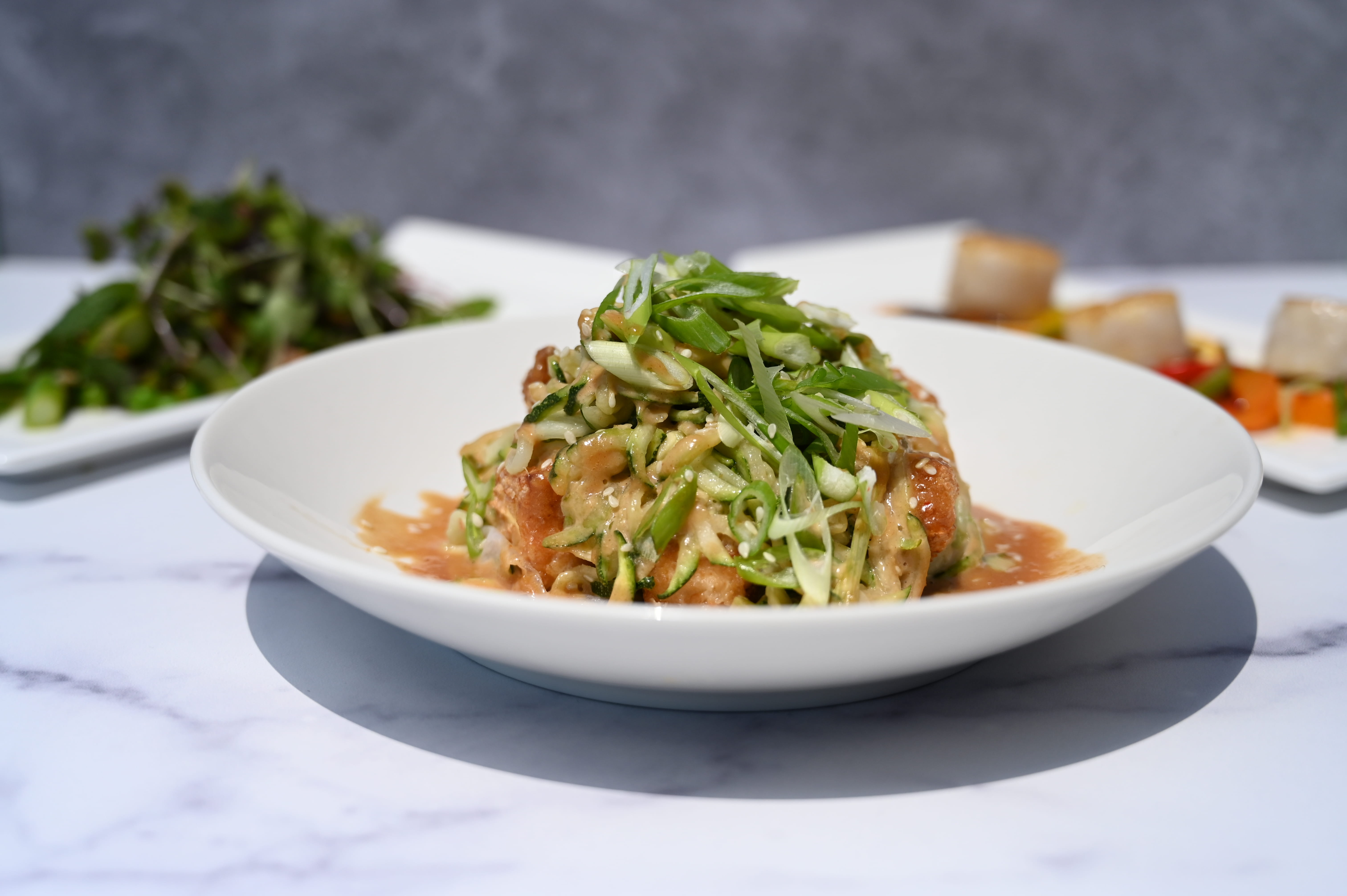 A colorful pad thai dish with scallions and noodles in front of a salad and a dish of pan seared scallops